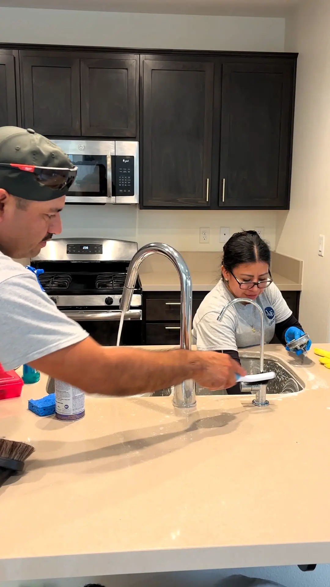 Two J&E Cleaning team members in uniform cleaning a kitchen sink and counter